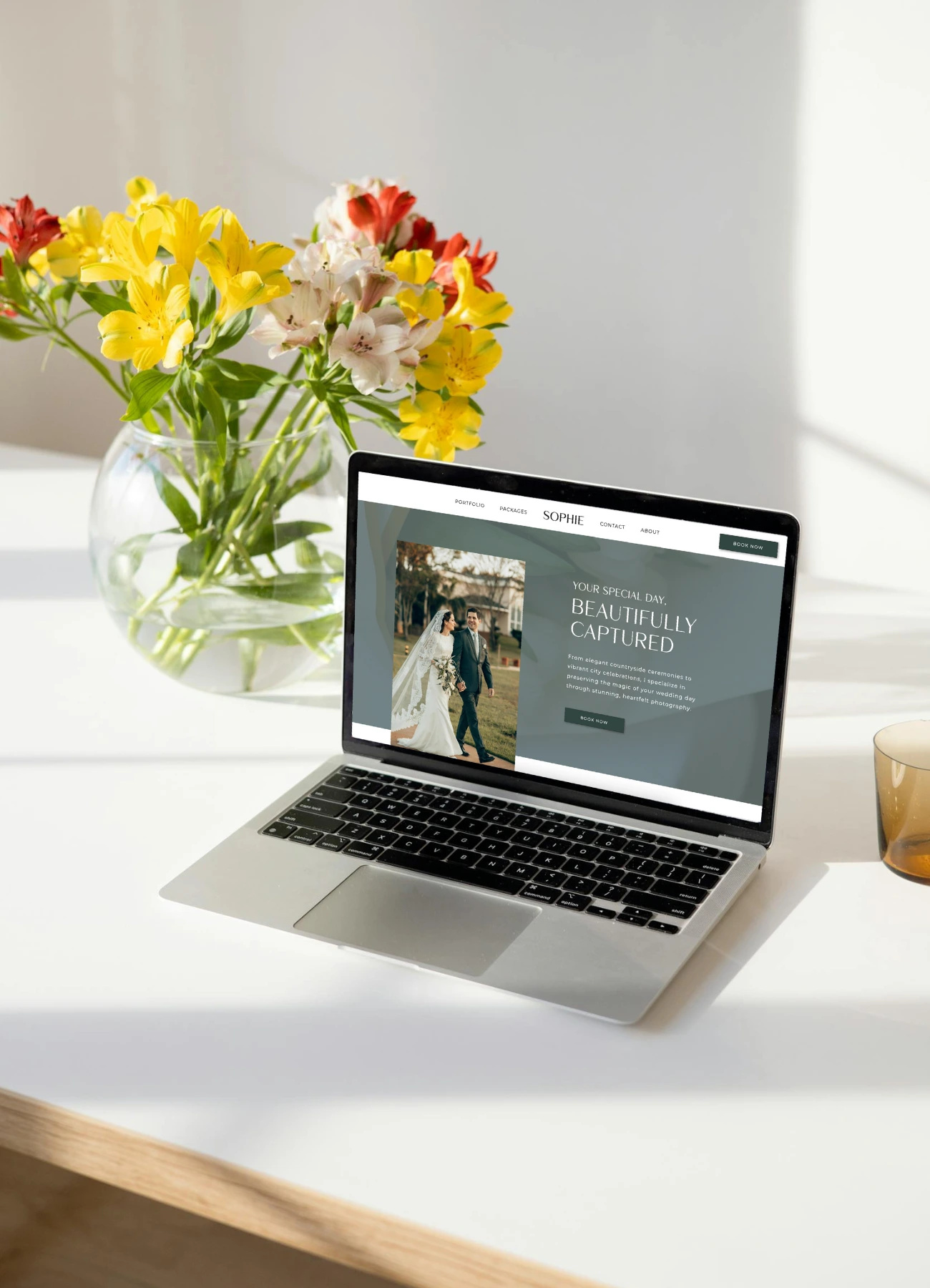 Laptop displaying a wedding photography website on a clean desk with flowers in the background.